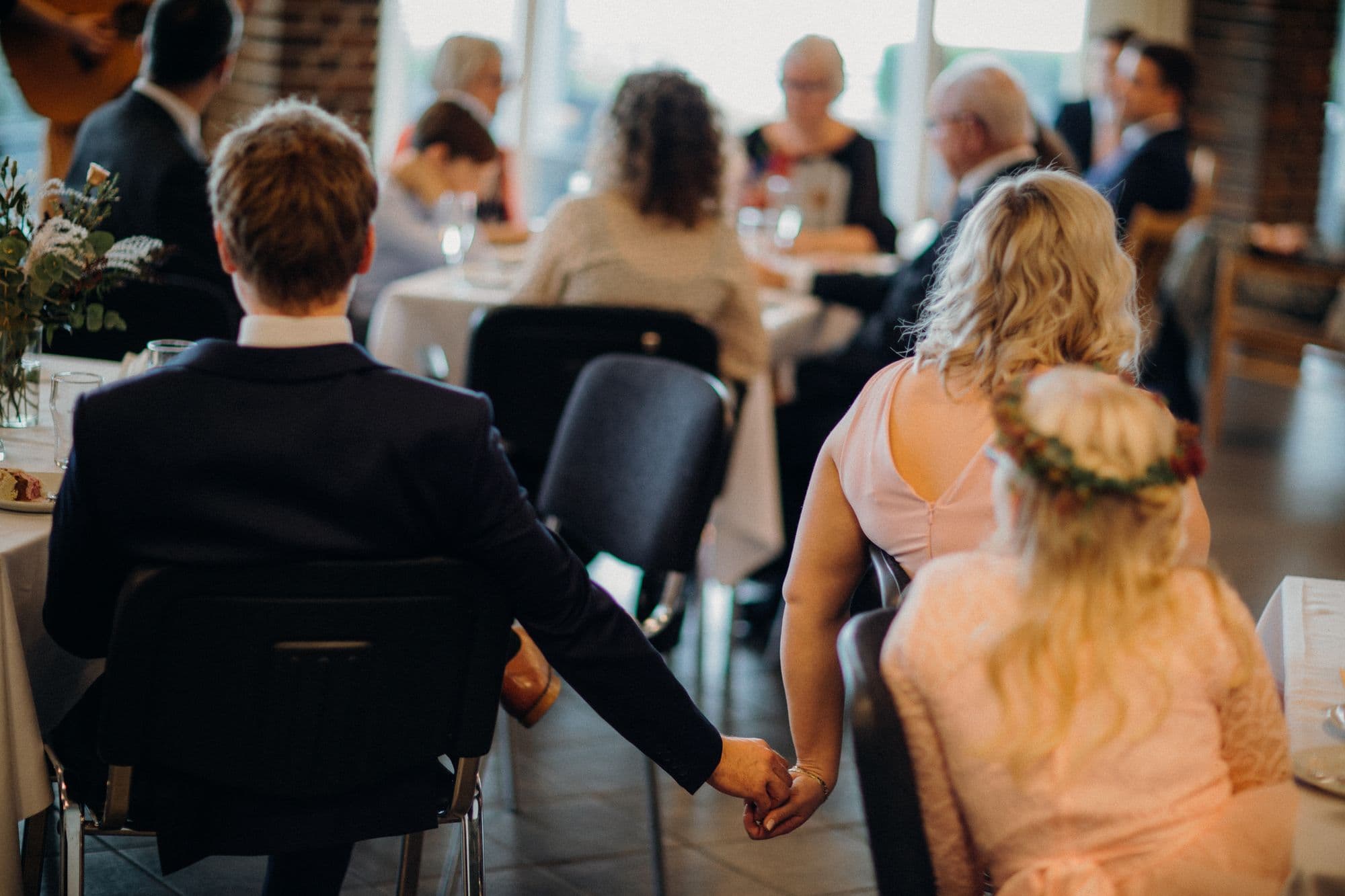 A couple holding hands from behind, guests filling a sunlit room around them
